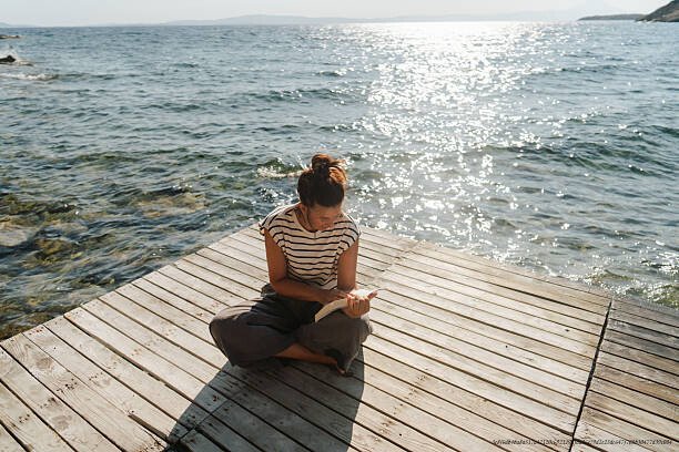 COMMENT RETOMBER À NOUVEAU AMOUREUX(E) APRÈS UNE DÉCEPTION SENTIMENTALE DOULOUREUSE EN 5 ÉTAPES 5 Photo of a mid adult woman reading a book and enjoy morning sun on the sea dock